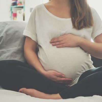 A heavily pregnant woman sits cross-legged with her arms protectively around the top and bottom of her belly.